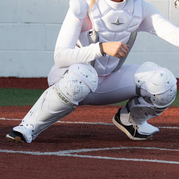 An All Star PHX Paige Halstead Fastpitch catcher in white protective gear crouches behind home plate, wearing All Star LGW-PHX leg guards with the LINQ hinge system for flexibility and Delta-Flex calf harness for a secure fit.