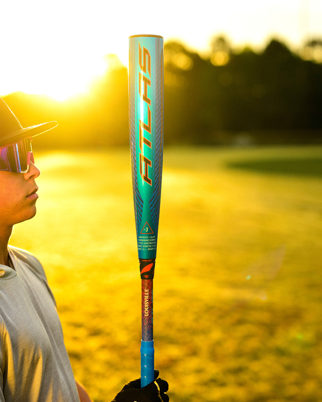 A person in sunglasses and a cap holds the 2027 Louisville Slugger Atlas Hybrid (-3) BBCOR Baseball Bat (WBL4246010) upright on a sunlit field, with trees softly blurred in the sunset background.