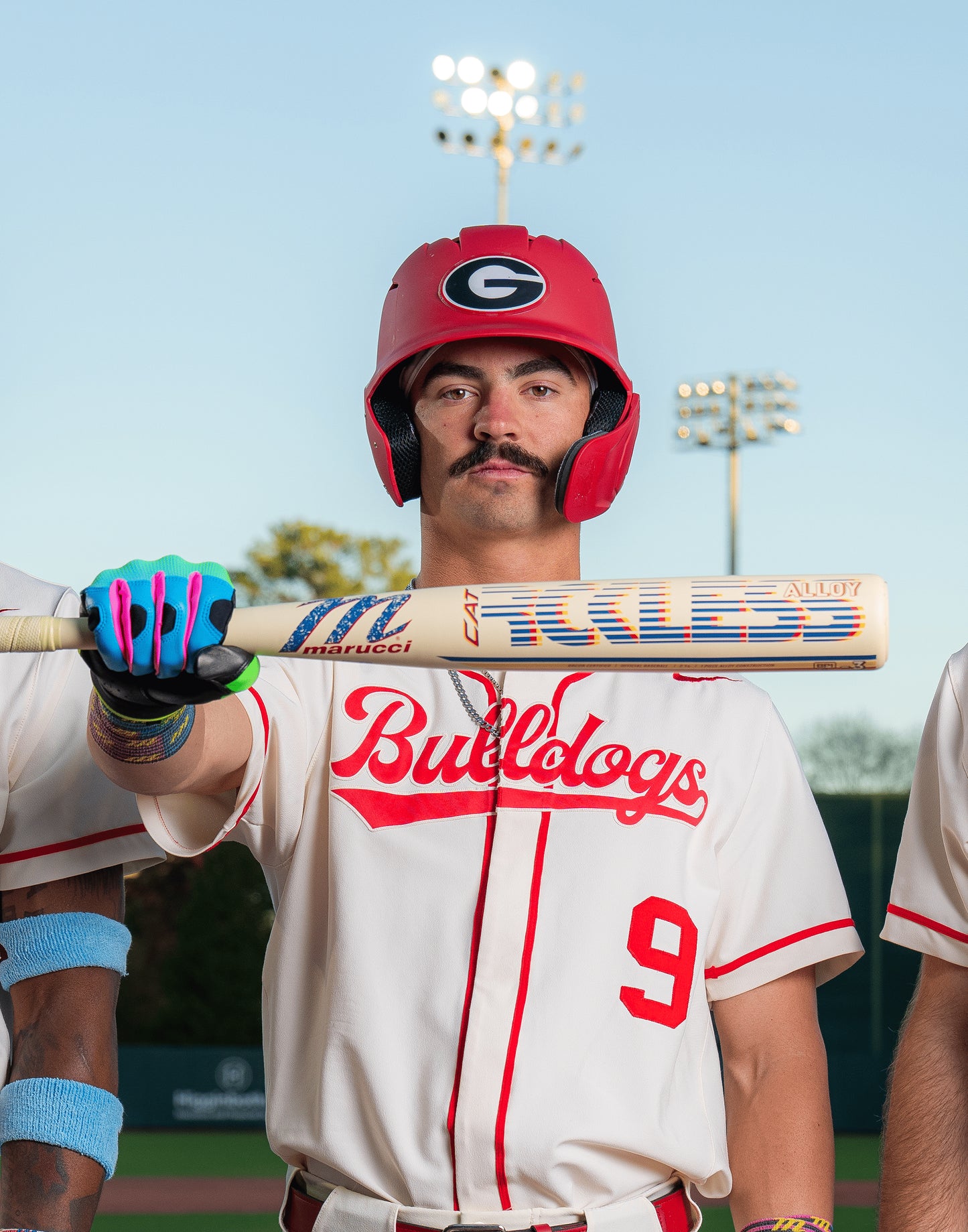 A baseball player in a red helmet and white Bulldogs #9 uniform holds the Marucci 2026 CATX RCKLESS Rewind (-3) BBCOR Baseball Bat (MCBCRRC) on a field, with stadium lights and two teammates partially visible.
