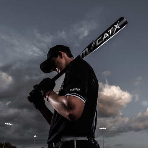 A baseball player in a black uniform and cap holds a bat over his shoulder, standing outdoors under a dramatic, cloudy sky with stadium lights in the background.