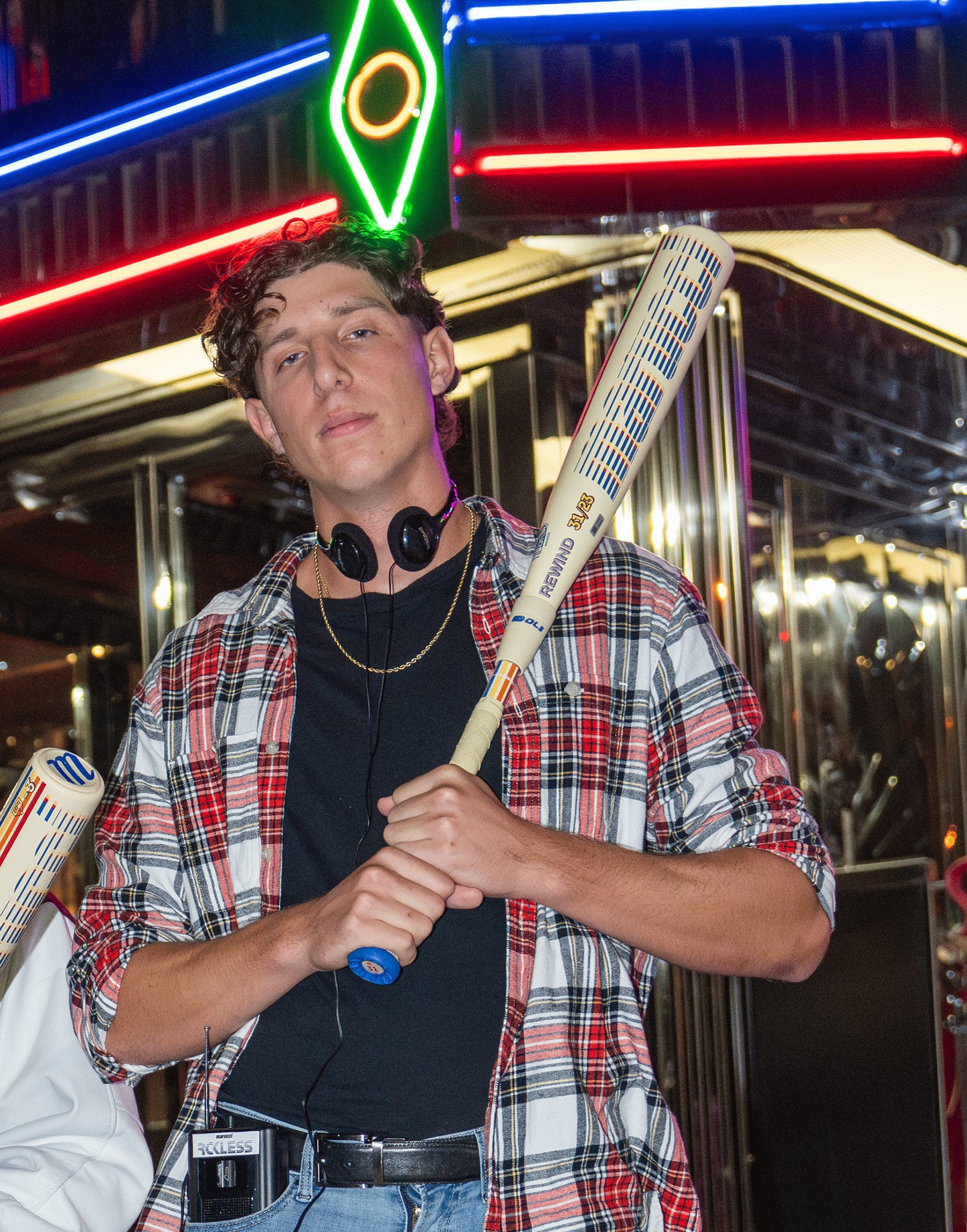 A young man in a plaid shirt with headphones around his neck holds a Marucci 2026 CATX RCKLESS Rewind Composite (-10) 2 3/4" USSSA Baseball Bat. Neon lights and metallic surfaces suggest a lively, nighttime urban scene.