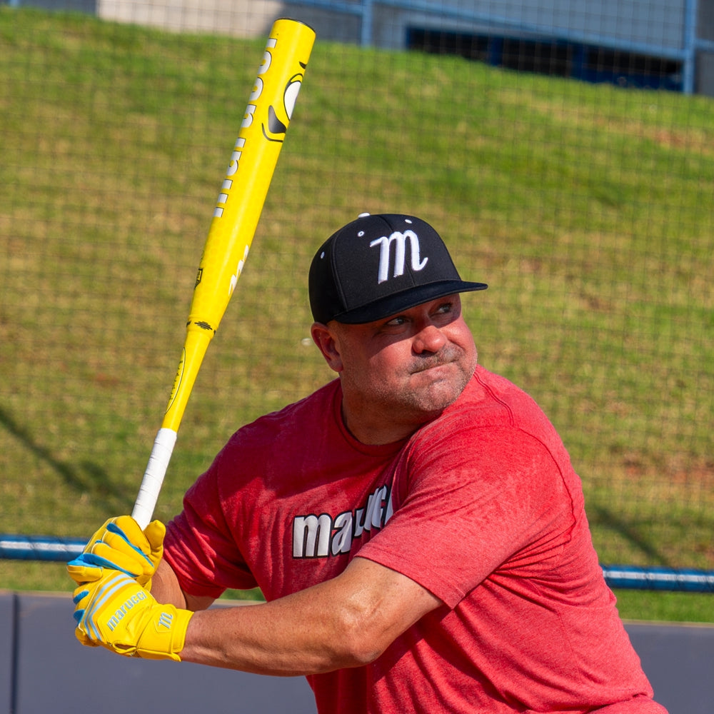 A man in a red shirt and black cap prepares to swing a yellow 2025 Marucci Chocolate Crush 12.75" Loaded USA Slowpitch Softball Bat (MSPCCY2LA). He wears yellow batting gloves, standing on grass with a wire fence behind him.