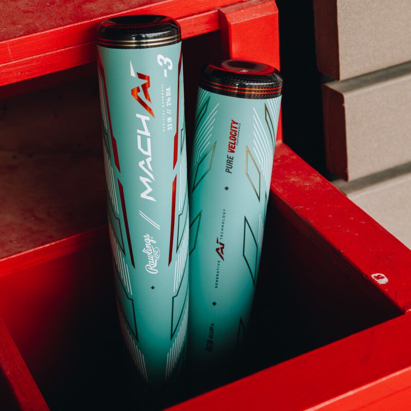 Two light blue Mach AI tennis ball cans with red and white graphics stand upright inside an open red container near a beige wall, next to a used 2024 Rawlings Mach AI (-3) BBCOR Baseball Bat: RBB4MC3.
