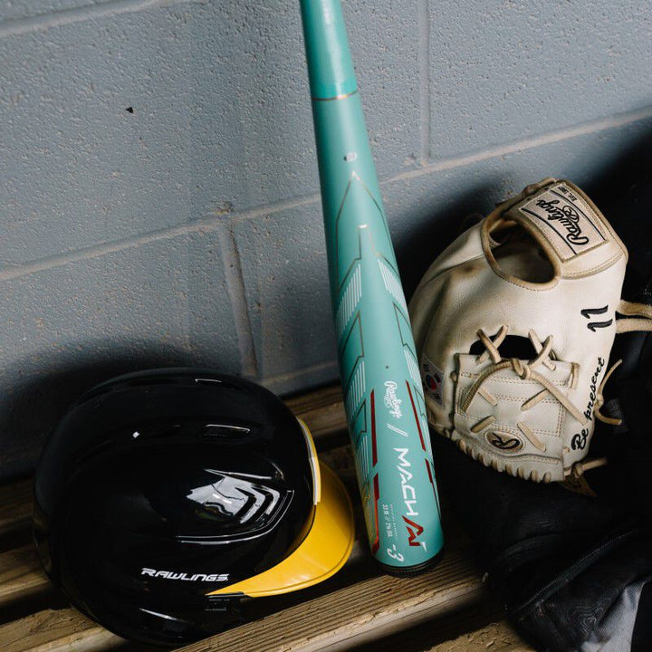 A 2024 Rawlings Mach AI (-3) BBCOR Baseball Bat (USED), a black Rawlings helmet, and a tan Rawlings glove rest on a wooden bench against a gray concrete wall.
