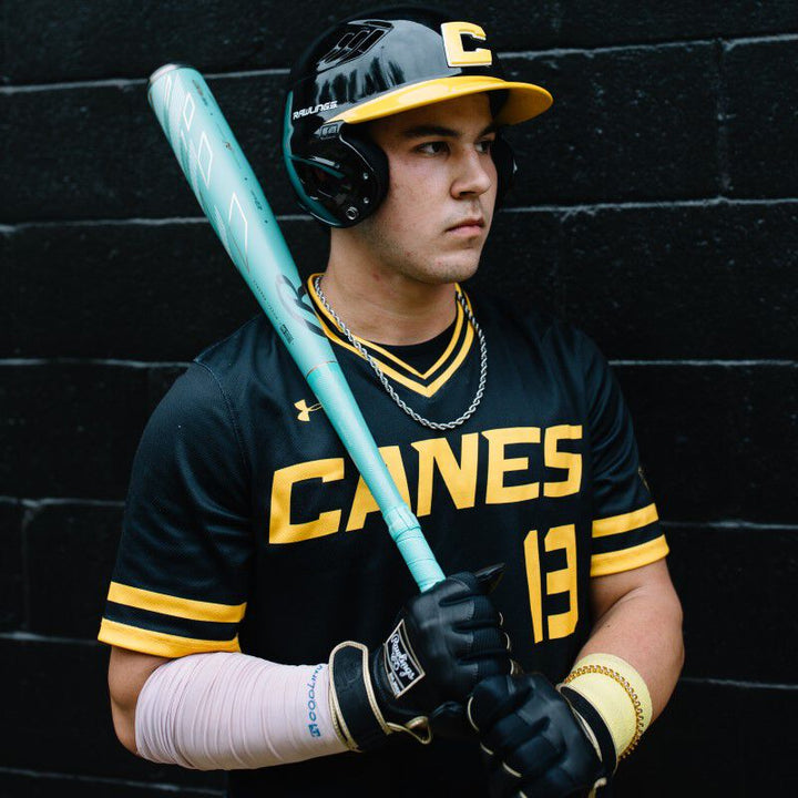 A baseball player in a black CANES uniform with yellow accents and #13 holds a used 2024 Rawlings Mach AI (-3) BBCOR Baseball Bat (RBB4MC3). He wears a black helmet, gloves, and has white tape on his left forearm, standing by a black brick wall.