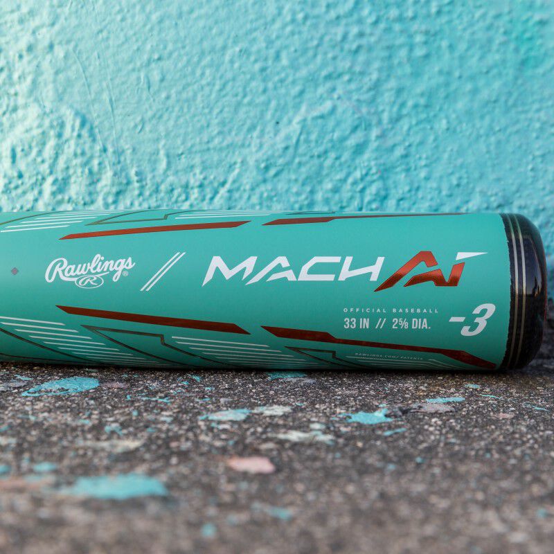 A used 2024 Rawlings Mach AI (-3) BBCOR Baseball Bat (RBB4MC3) in teal with red and white lettering rests on worn concrete against a turquoise wall. The alloy bat features a 33" length, 2 5/8" barrel, and -3 drop.