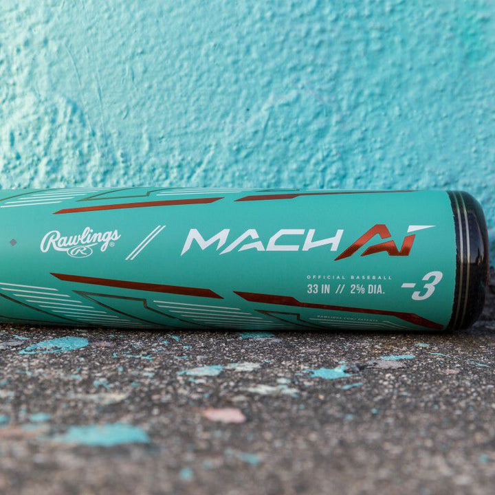 A used 2024 Rawlings Mach AI (-3) BBCOR Baseball Bat (RBB4MC3) in teal with red and white lettering rests on worn concrete against a turquoise wall. The alloy bat features a 33" length, 2 5/8" barrel, and -3 drop.