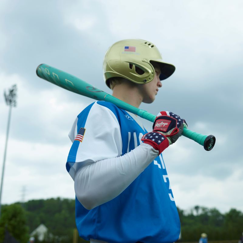 A baseball player in a gold helmet and blue jersey holds a used 2024 Rawlings Mach AI (-3) BBCOR Baseball Bat over his shoulder. He wears American flag gloves, with trees and a cloudy sky in the background.