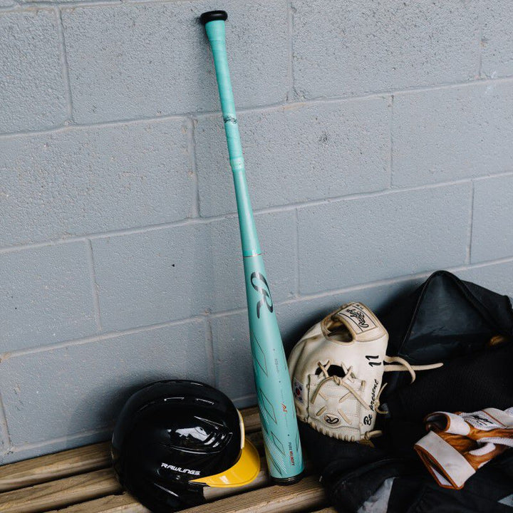 A 2024 Rawlings Mach AI (-3) BBCOR Baseball Bat (USED) by Rawlings leans against the wall alongside a helmet, poised for the next big game.