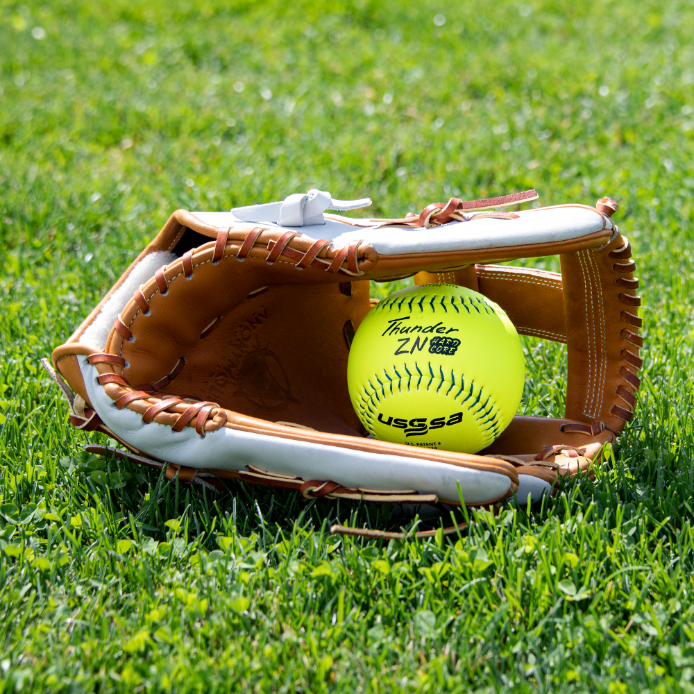 A Dudley USSSA Thunder ZN Hard Core Pro W 11" 44/375 Composite Slowpitch Softball (4U11H) sits in an open leather baseball glove on green grass under bright sunlight.