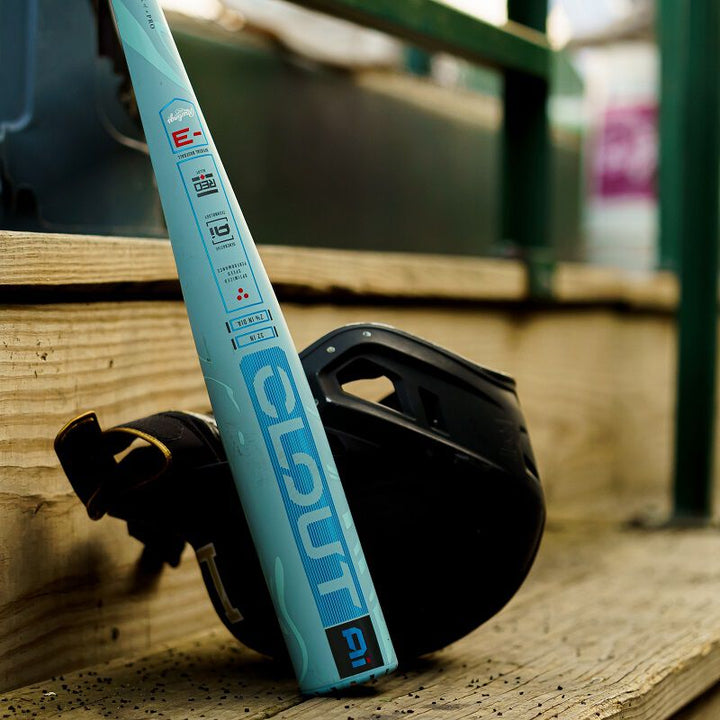 A blue 2025 Rawlings Clout AI (-3) BBCOR Baseball Bat: RBB5C3 (USED) rests against wooden bleachers next to a black helmet, with green railings in the blurred background.