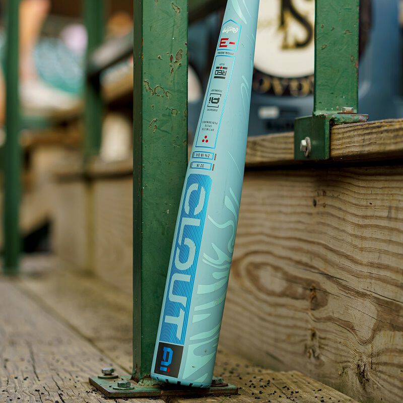 A close-up of the 2025 Rawlings Clout AI (-3) BBCOR Baseball Bat (USED), a light blue Rawlings bat with bold blue lettering, leaning against a green metal railing on a wooden platform, with a blurred background.