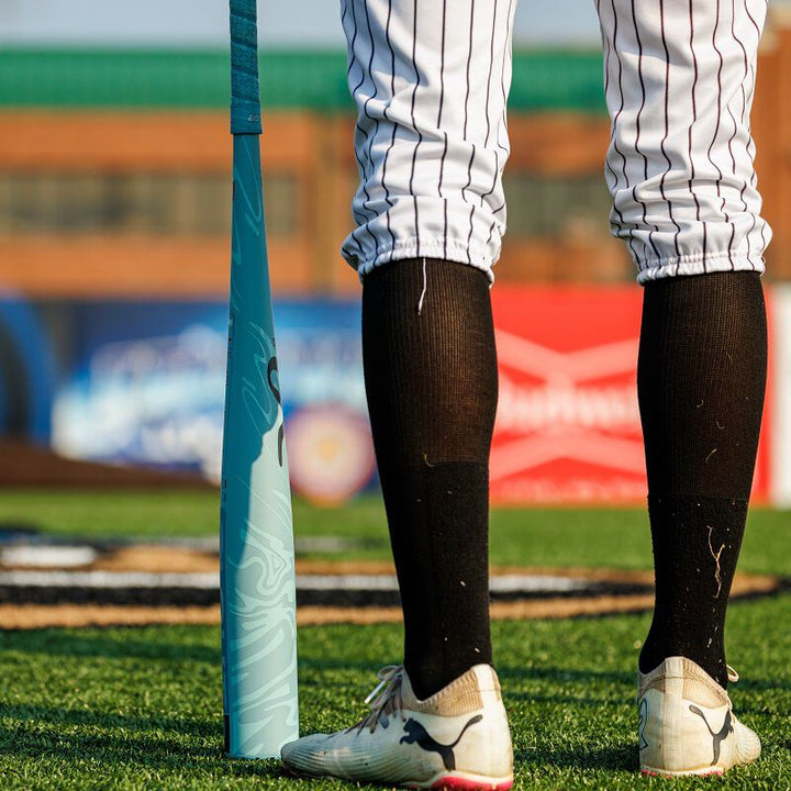 A baseball player in white cleats, black socks, and pinstriped pants stands on a field next to a 2025 Rawlings Clout AI (-3) BBCOR Baseball Bat: RBB5C3 (USED), with blurred stadium signage in the background.