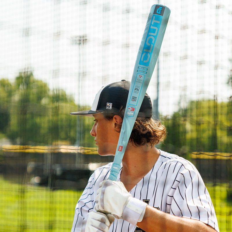 A baseball player in a pinstripe jersey and black cap holds a 2025 Rawlings Clout AI (-3) BBCOR Baseball Bat: RBB5C3 (USED) over his shoulder at home plate, looking to the side. The background features a field and protective netting.