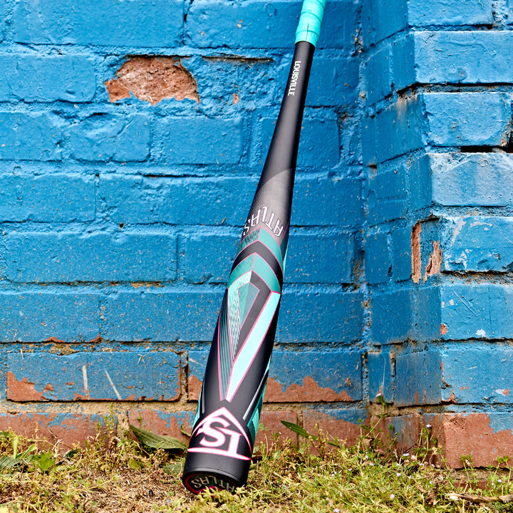 A 2025 Louisville Slugger Atlas (-3) BBCOR Baseball Bat (WBL2968010 DEMO), black and teal with geometric designs, leans against a weathered blue brick wall on grass and dirt.