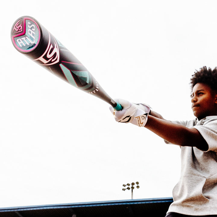 A young person in a gray shirt and white gloves holds the 2025 Louisville Slugger Atlas (-3) BBCOR Baseball Bat: WBL2968010 (DEMO) toward the camera, with a stadium and lights shining in the background.