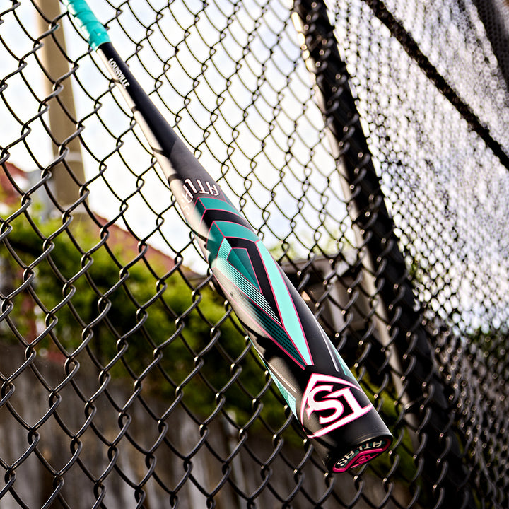 A 2025 Louisville Slugger Atlas (-3) BBCOR Baseball Bat (WBL2968010, DEMO) hangs diagonally against a chain-link fence, with a blurred background of trees and a wooden fence in daylight.