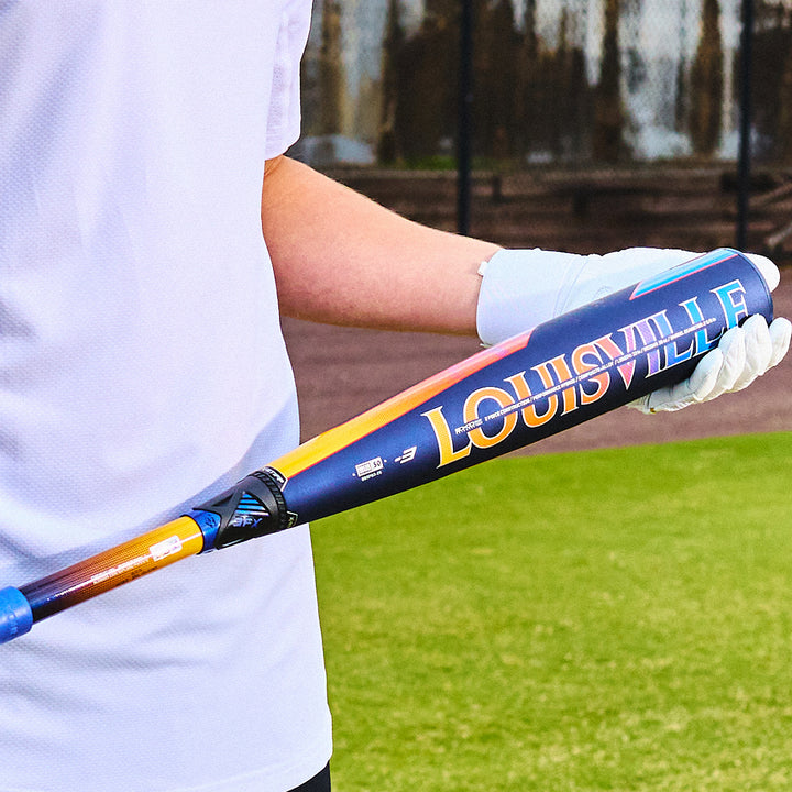 A person in a white shirt and glove holds a colorful 2025 Louisville Slugger Select PWR (-3) BBCOR Baseball Bat: WBL2967010 (DEMO) on a grassy field, with a fence and dirt visible in the background.