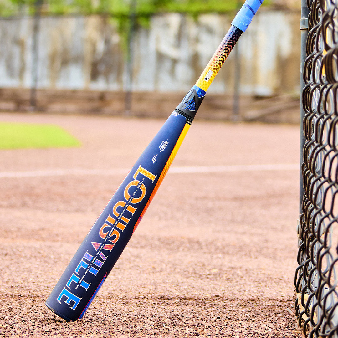 A 2025 Louisville Slugger Select PWR (-3) BBCOR Baseball Bat (WBL2967010 DEMO) stands upright against a chain-link fence on a baseball field, with grass and the dirt infield in the background.