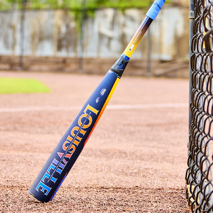 A 2025 Louisville Slugger Select PWR (-3) BBCOR Baseball Bat (WBL2967010 DEMO) stands upright against a chain-link fence on a baseball field, with grass and the dirt infield in the background.