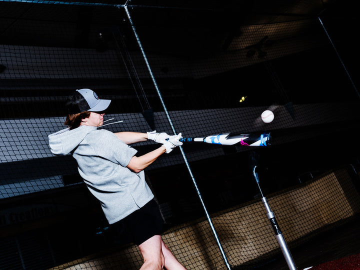 Wearing a gray hoodie and cap, a batter hits a baseball with a 2025 DeMarini ZEN (-10) 2 3/4" USSSA Bat (WBD2532010, USED) inside a batting cage. The ball is lit midair against the dark background.