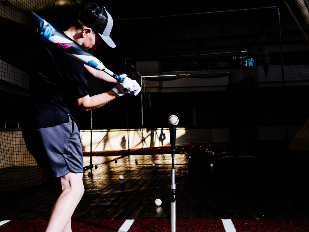A young player in a cap and gloves swings a used DeMarini 2025 ZEN (-10) 2 3/4" USSSA Baseball Bat (WBD2532010) at a ball on a tee in a dim indoor batting cage, with scattered baseballs on the floor.