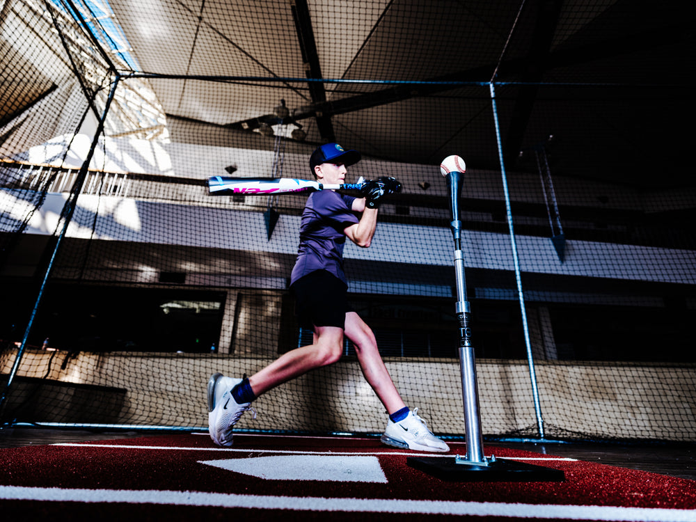 A young baseball player in athletic gear swings a 2025 DeMarini ZEN (-10) 2 3/4" USSSA Baseball Bat (USED) at a ball on a tee inside an indoor practice facility, with sunlight streaming through the windows.