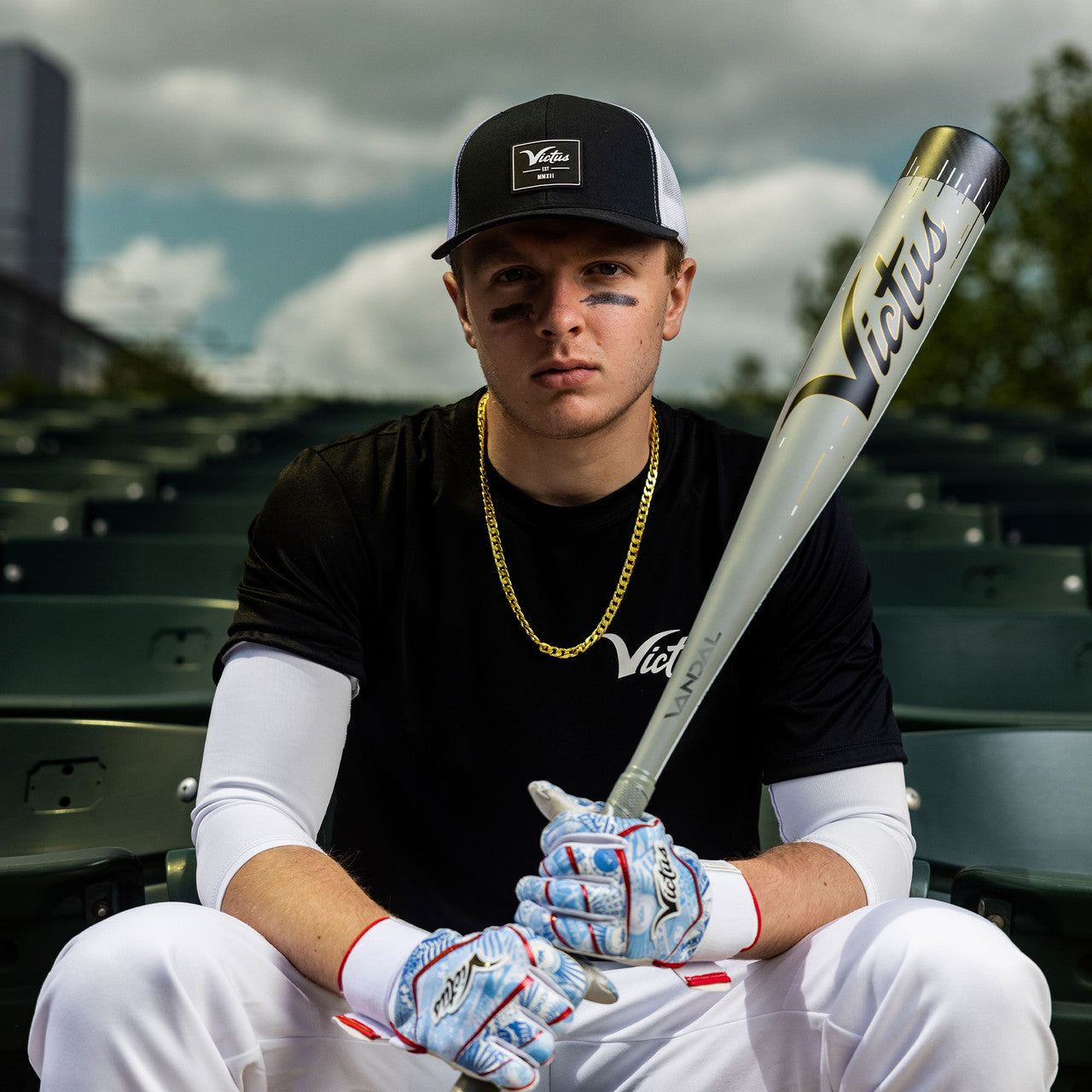 A baseball player in a black cap and shirt, white pants, and gold chain sits in stadium seats holding a USED 2024 Victus Vandal Lev3 (-3) BBCOR Baseball Bat (VCBV3). He wears batting gloves and eye black, staring seriously at the camera.