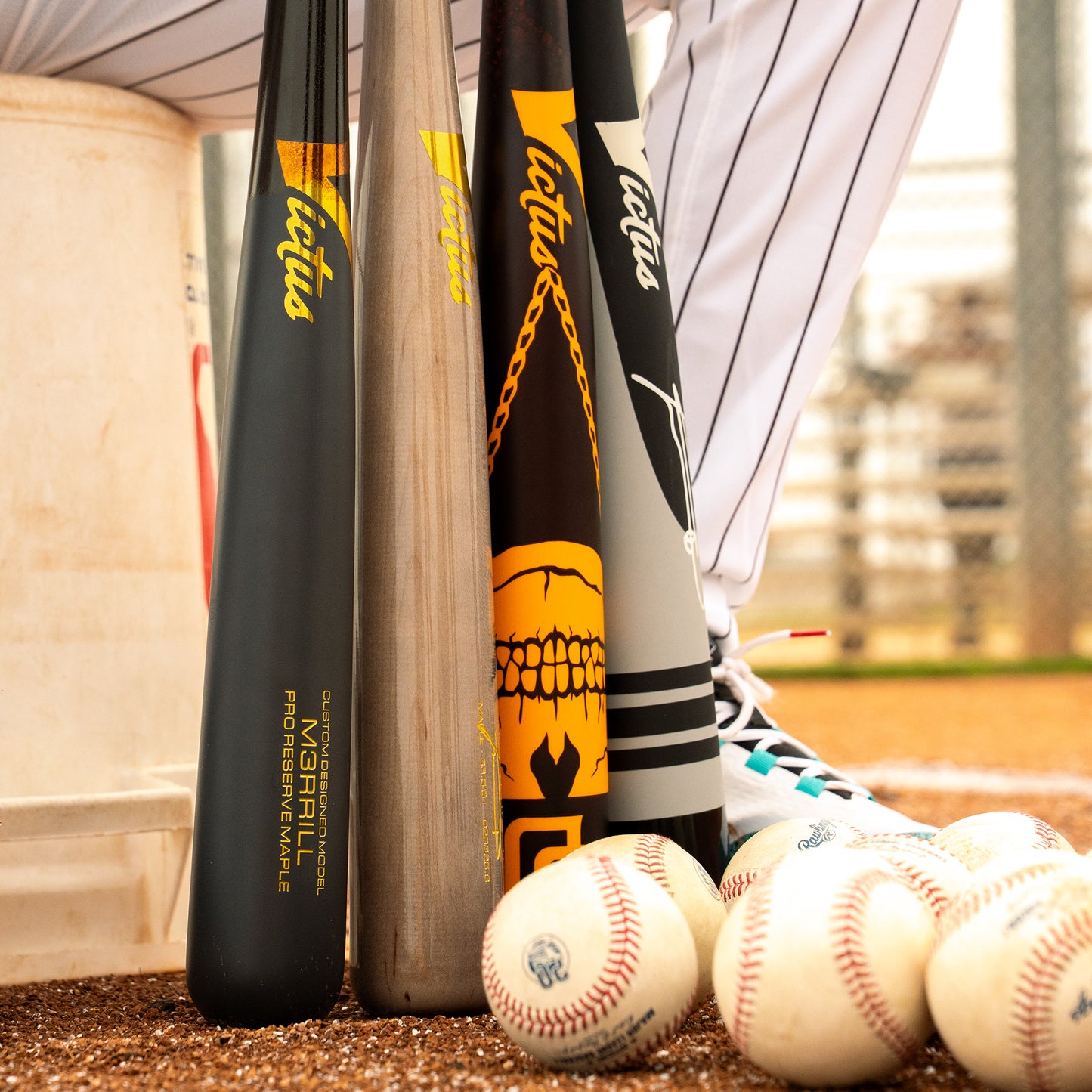 Three Victus Jackson Merrill M3RRILL Pro Reserve Maple Wood Bats by Victus stand upright on a field next to a player’s leg in striped pants, with baseballs scattered around and part of a white bucket visible on the left.