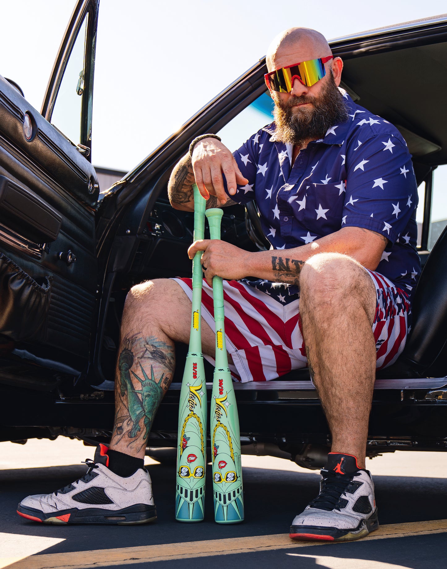 A bearded man in a star-patterned shirt, striped shorts, and colorful sunglasses sits in a car with the door open, holding two Victus Nova Lit Beacon (-8) baseball bats (VSBNL8B), showing off his tattoos and sneakers.
