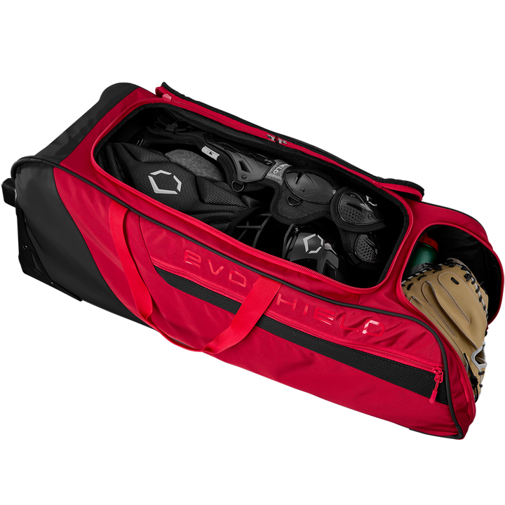 The EvoShield Takeover Wheeled Bag (WB57572) by EvoShield is unzipped, showing spacious bat sleeves and baseball equipment inside, including a helmet, catcher’s gear, and a tan glove.