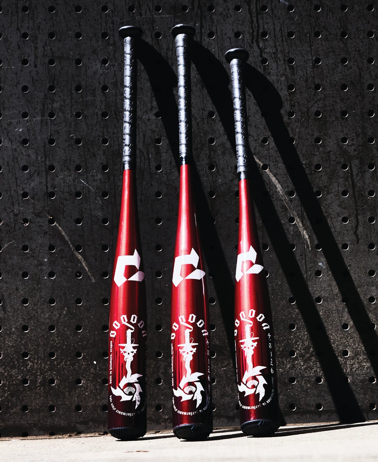 Three 2025 DeMarini Voodoo One (-8) USSSA baseball bats (WBD2539010) by DeMarini, featuring a red and black design with white graphics, stand upright against a dark perforated wall, casting distinct shadows.