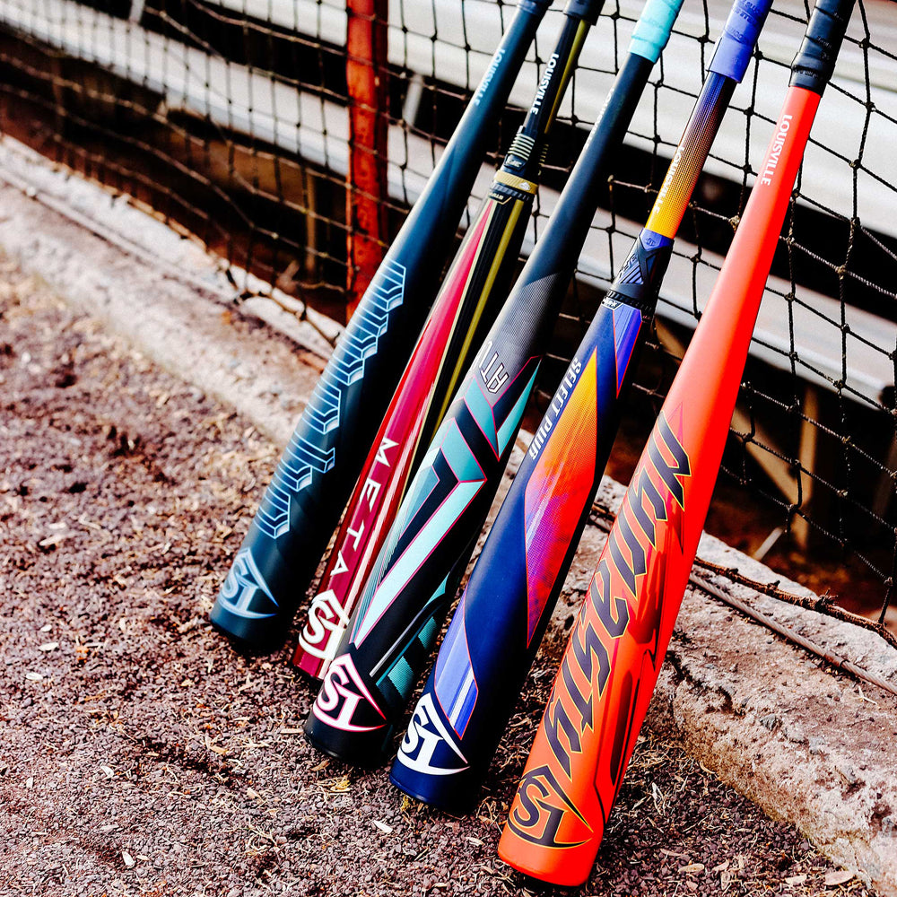 Four colorful baseball bats, including a 2025 Louisville Slugger Dynasty (-3) BBCOR Baseball Bat: WBL2969010, are lined up upright against a metal fence on dirt, each with its handle resting on the fence.