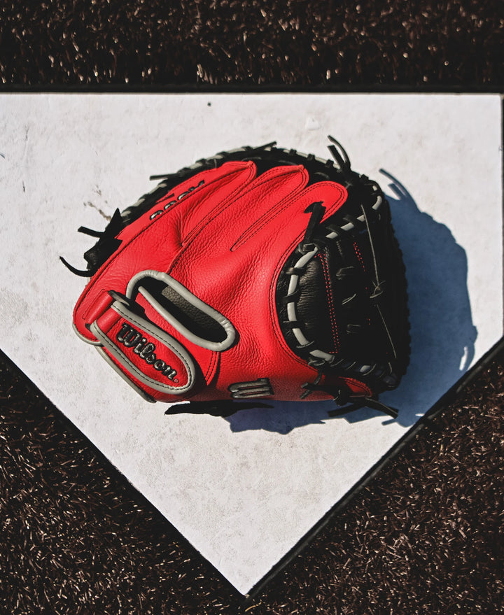 A Wilson A500 32" Baseball Catcher's Mitt (WBW10254632) by Wilson rests on home plate, its red color casting a shadow across the white surface and standing out against the dark brown dirt around the plate.