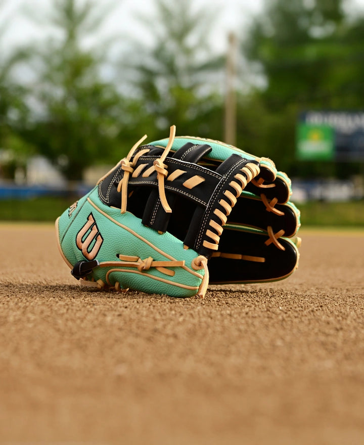 A Wilson A2000 1716SS 11.5" Baseball Glove (SPRING 2025: WBW102547115) in teal and black with tan laces rests on a baseball field's dirt, with trees and a blurred scoreboard seen in the background.