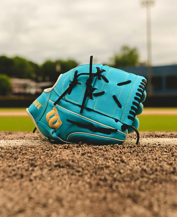 A Wilson A2000 SA17 12" Baseball Glove (SPRING 2025: WBW10255012) in teal with black laces sits on a pitcher’s mound, outfield and trees softly blurred in the background.