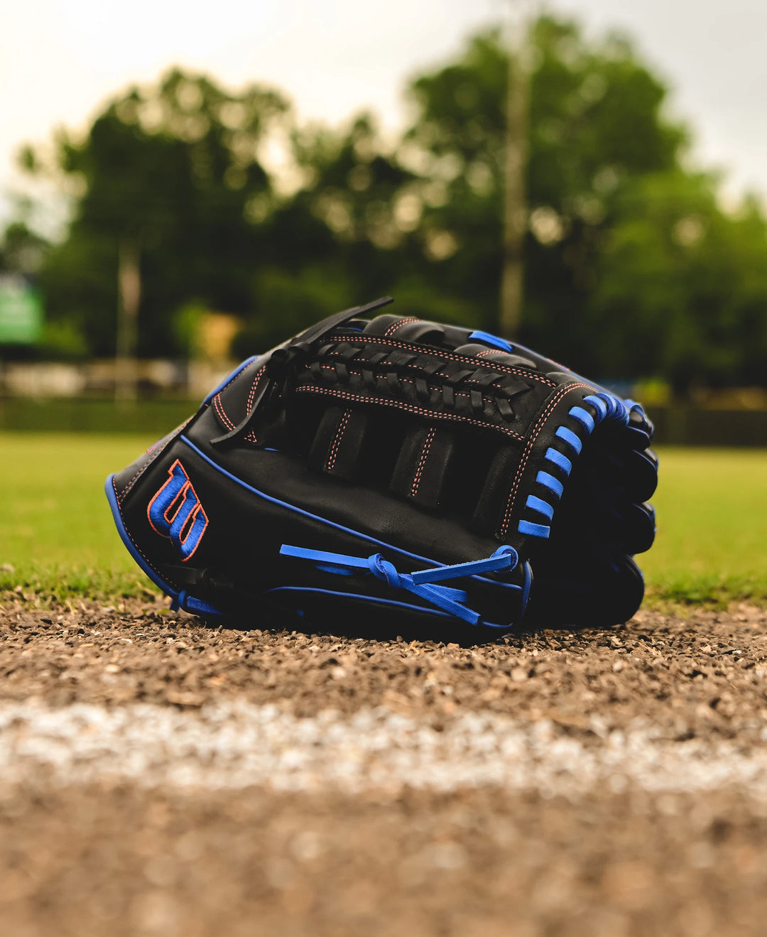 A Wilson A1000 PF1892 12.25" Baseball Glove (WBW1025831225) rests on the ground near the edge of a baseball field, framed by green trees and a softly blurred background.