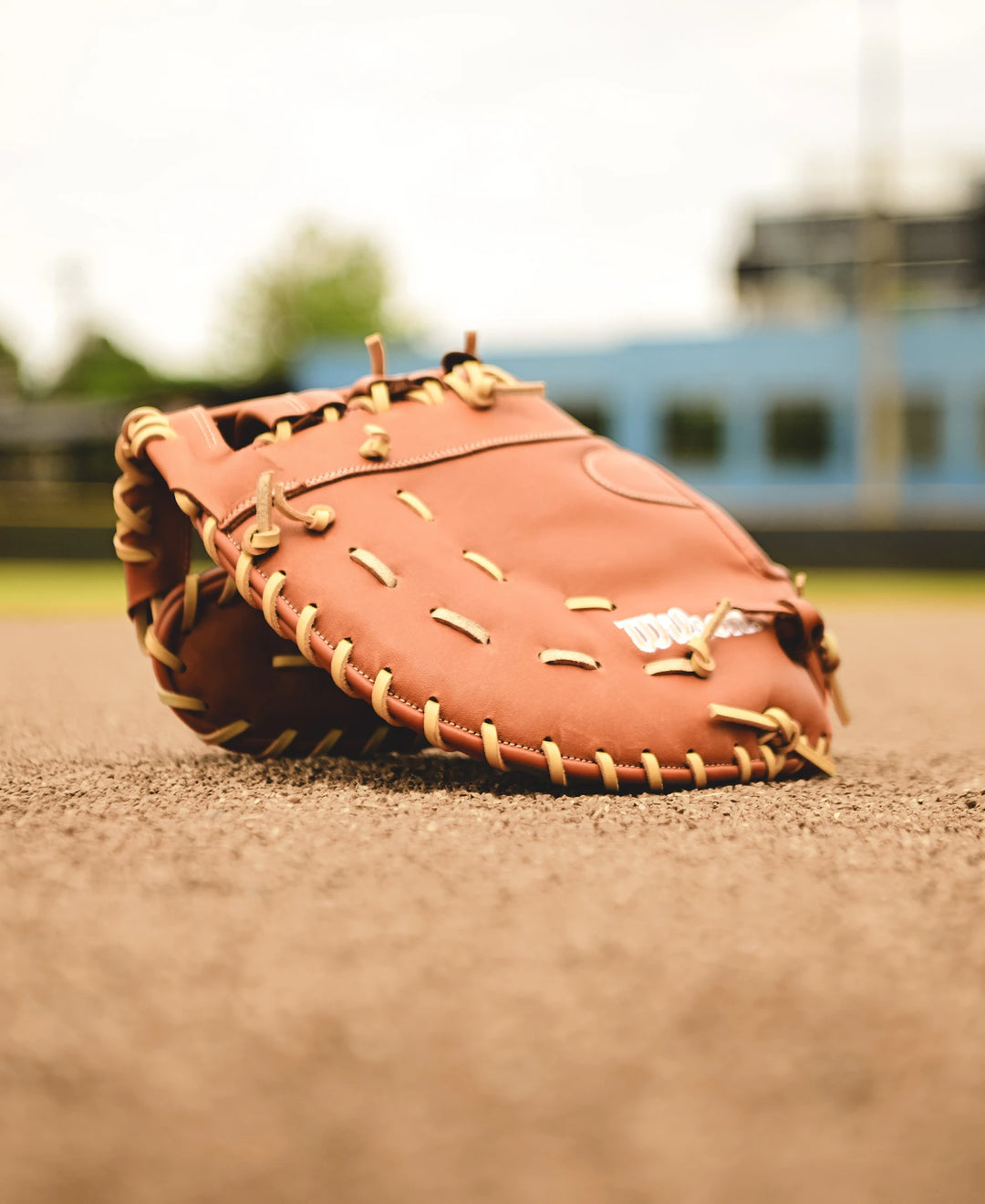 A Wilson A1000 1620 12.5" Baseball First Base Mitt (WBW102587125) made from full grain leather lies on the infield dirt, with blurred green trees and blue buildings in the background.
