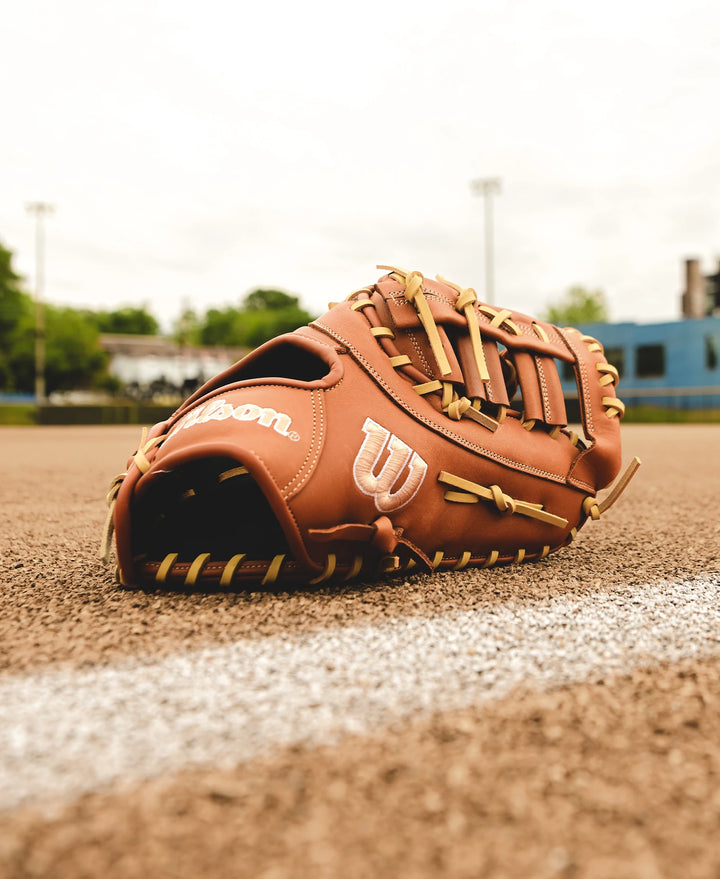A Wilson A1000 1620 12.5" First Base Mitt (WBW102587125) from Wilson lies on the dirt near a chalk line on a baseball field, with green trees and blurred stadium lights in the background.