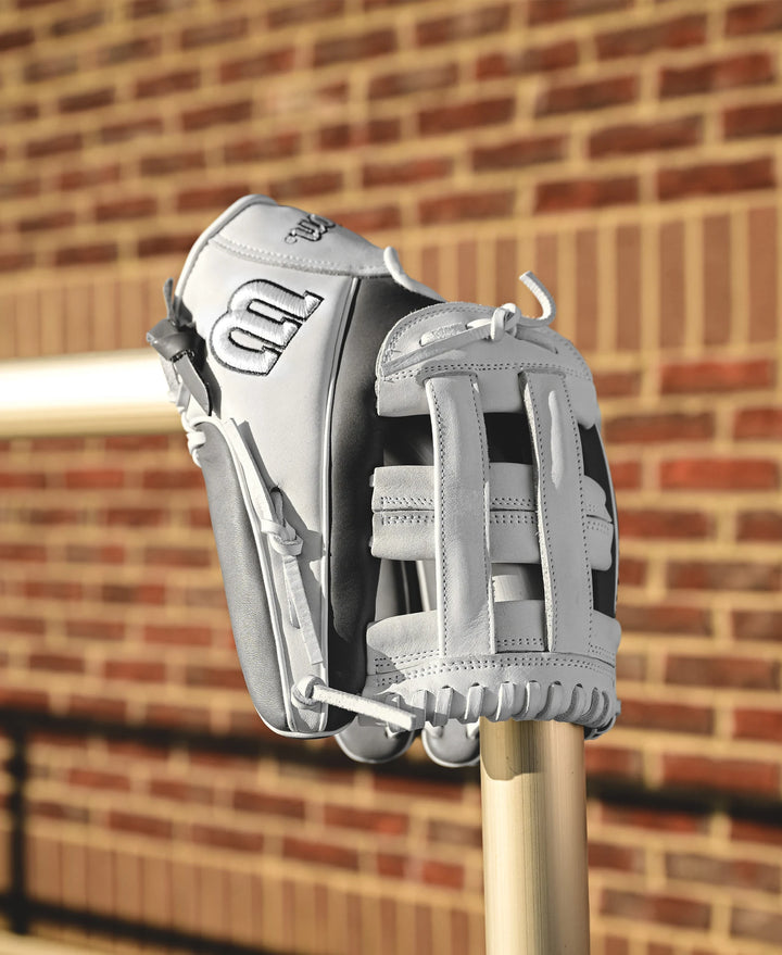 A Wilson A1000 IF12 12" Fastpitch Glove by Wilson in white and gray full grain leather sits atop a metal pole, with a blurred brick wall and railing in the background.