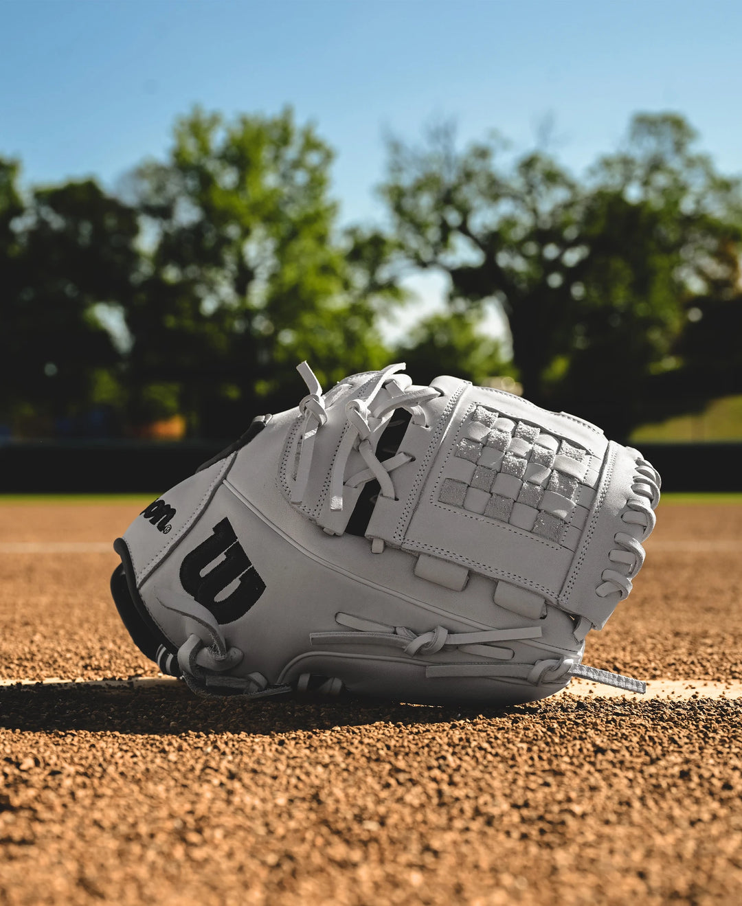 A Wilson A1000 P12 12" Fastpitch Glove (WBW10259212) rests on a dirt infield, framed by green trees and a blurred fence beneath a clear blue sky.