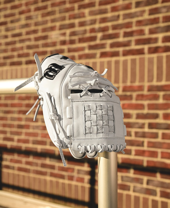 A Wilson A1000 P12 12" Fastpitch Glove (WBW10259212), made of white full grain leather with black accents, sits on a metal rail with a brick wall in the background.