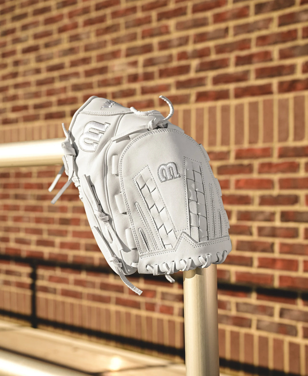 The Wilson A1000 V125 12.5" Fastpitch Glove (WBW102594125) by Wilson sits on a metal railing with a brick wall behind it, bathed in sunlight and casting shadows.