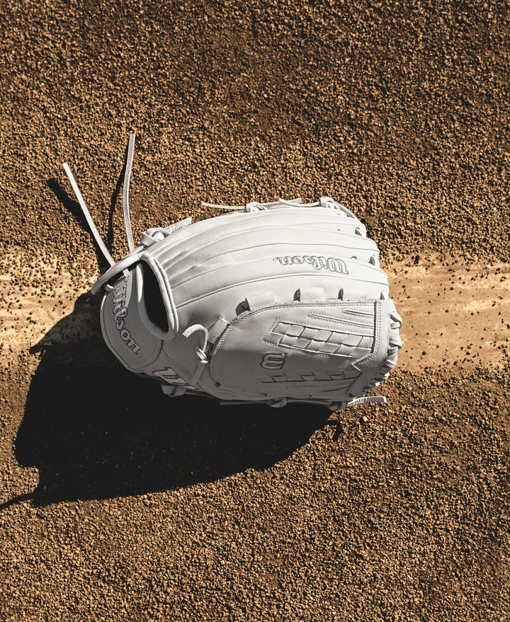 A Wilson A1000 V125 12.5" Fastpitch Glove (WBW102594125) rests on dirt, partly covering a bright white foul line, casting a shadow to the left on the textured field.