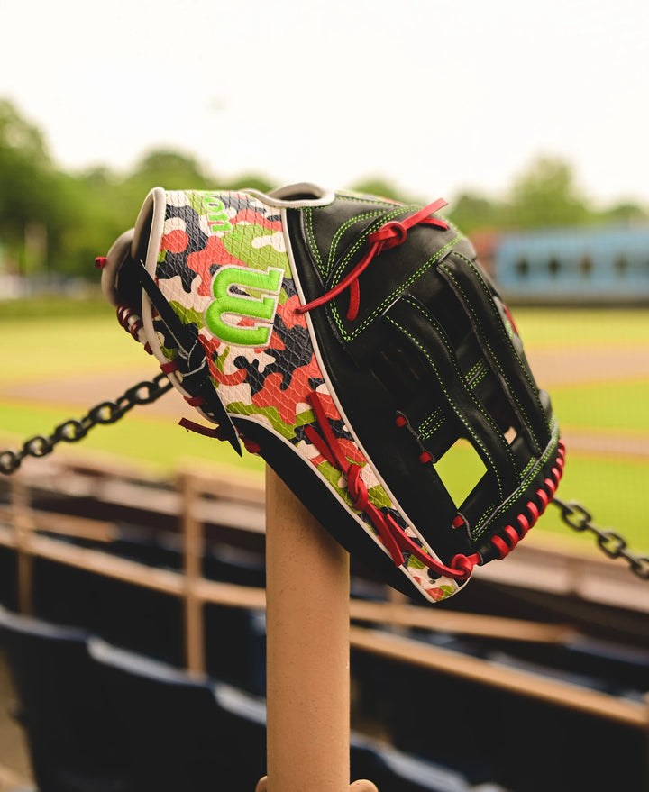 A Wilson A2K MH23 12.75" Michael Harris GM Baseball Glove (WBW1026201275) with a camouflage pattern and red laces sits on a bat, crafted from Pro Stock Select leather, against a blurred empty field and bleachers.