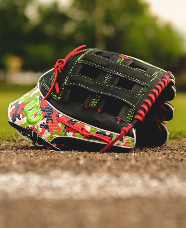 A Wilson A2K MH23 12.75" Michael Harris GM Baseball Glove, featuring red laces and camouflage patterns, rests near the edge of a dirt baseball field with green grass and blurred trees in the background.