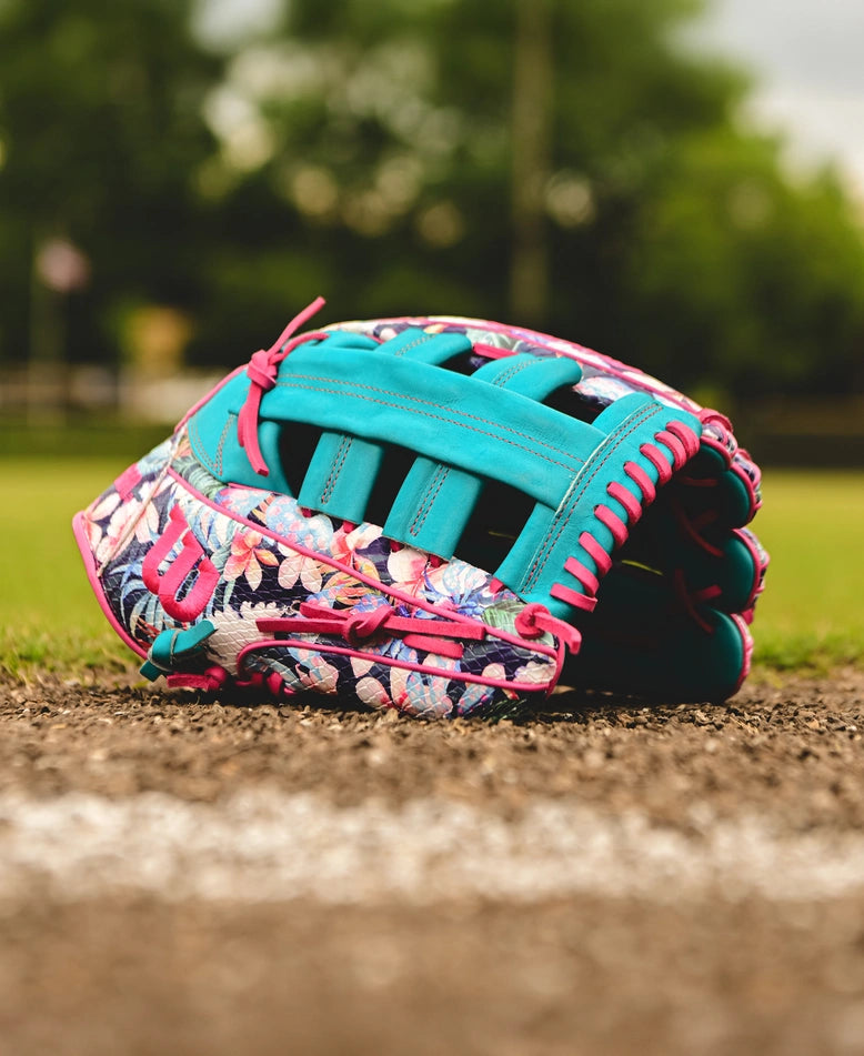 A Wilson A2000 RA56 12.75" Randy Arozarena GM Baseball Glove featuring blue, pink, and floral patterns lies on the dirt by a white baseline, with blurred grass and trees in the background.