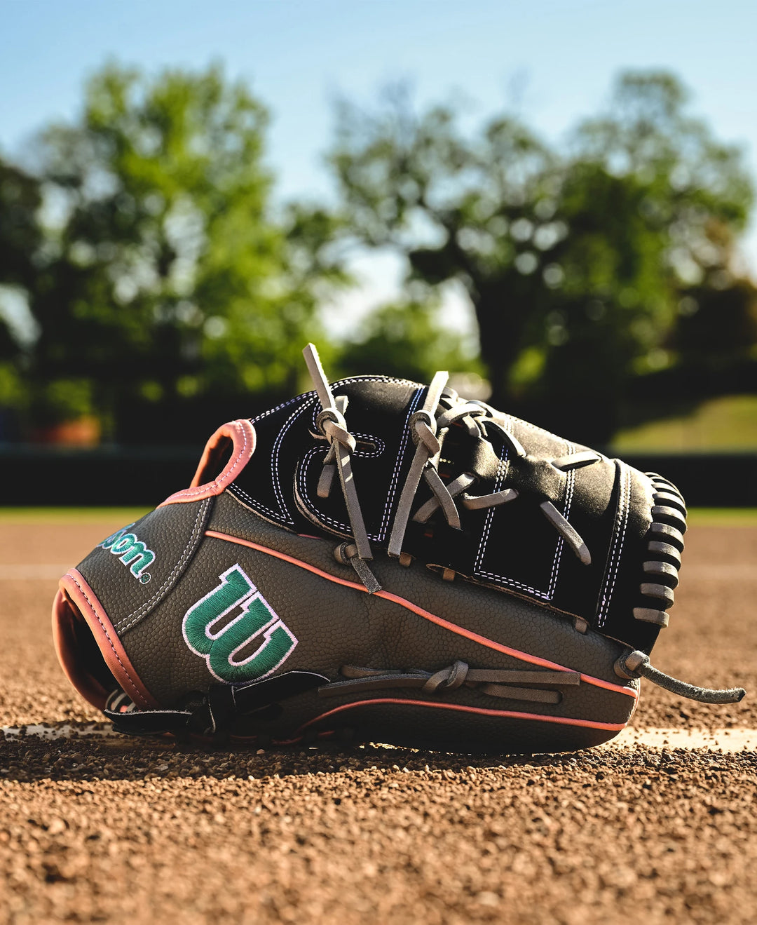 A close-up of the Wilson A2000 MA14SS 12.25" Fastpitch Glove (WBW1027221225) lies on a dirt baseball field, with green trees and a clear blue sky softly blurred in the background.