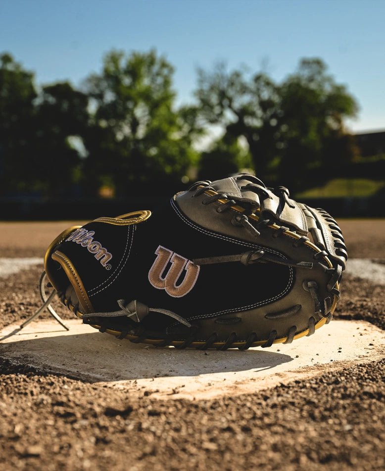 A Wilson A2000 FPCM23 34" Fastpitch Catcher's Mitt (SPRING 2025: WBW10274434) sits on the pitcher’s mound under a clear blue sky, surrounded by green trees and a blurred field in the background.