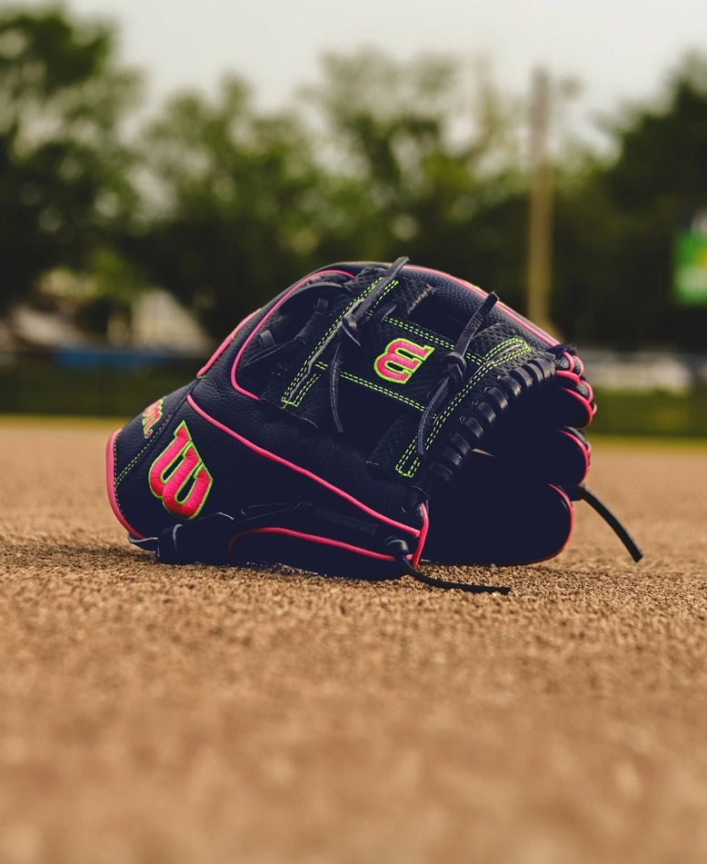 The Wilson A2000 DP15SS 11.5" Baseball Glove (SPRING 2025: WBW102747115) with green and pink accents rests on the infield dirt, featuring Pedroia Fit Technology, with blurred trees and field equipment in the background.